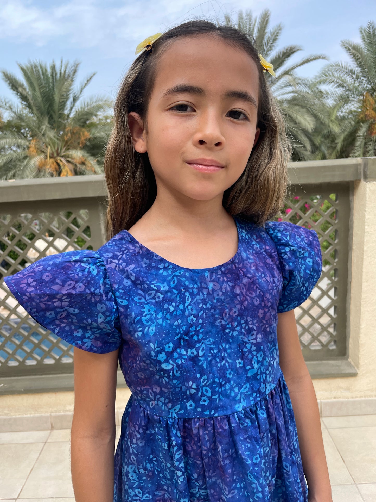 Front close up view of girl wearing a cotton batik dress in a contrasting blue floral print, showing the ruffle sleeves and bold floral print, sustainably handmade in Bali, for sale in Dubai.