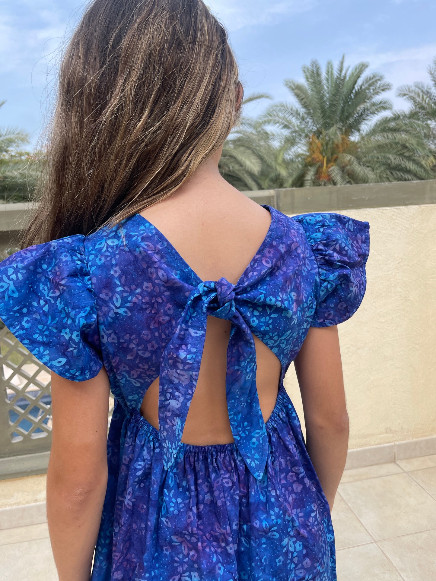 Back close up view of girl wearing a cotton batik dress in a contrasting blue floral print, showing the ruffle sleeves and adjustable back ties, sustainably handmade in Bali, for sale in Dubai.