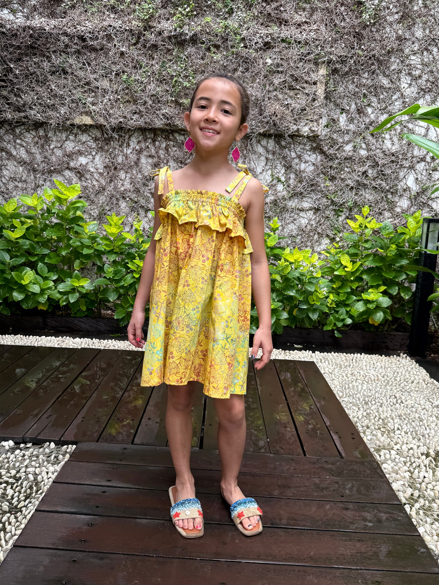 Front pose of girl wearing mustard yellow floral batik cotton sundress with adjustable shoulder ties and ruffles, sustainably made in Bali, available in Dubai