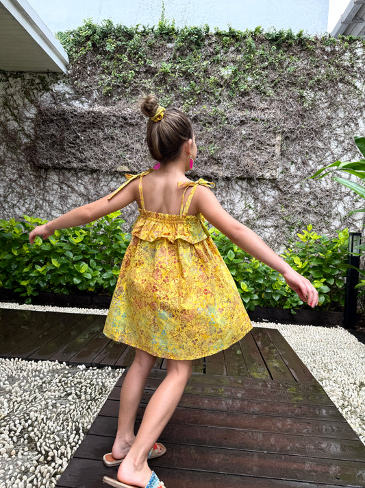 Girl twirling and wearing mustard yellow floral batik cotton sundress with adjustable shoulder ties and ruffles in a back pose