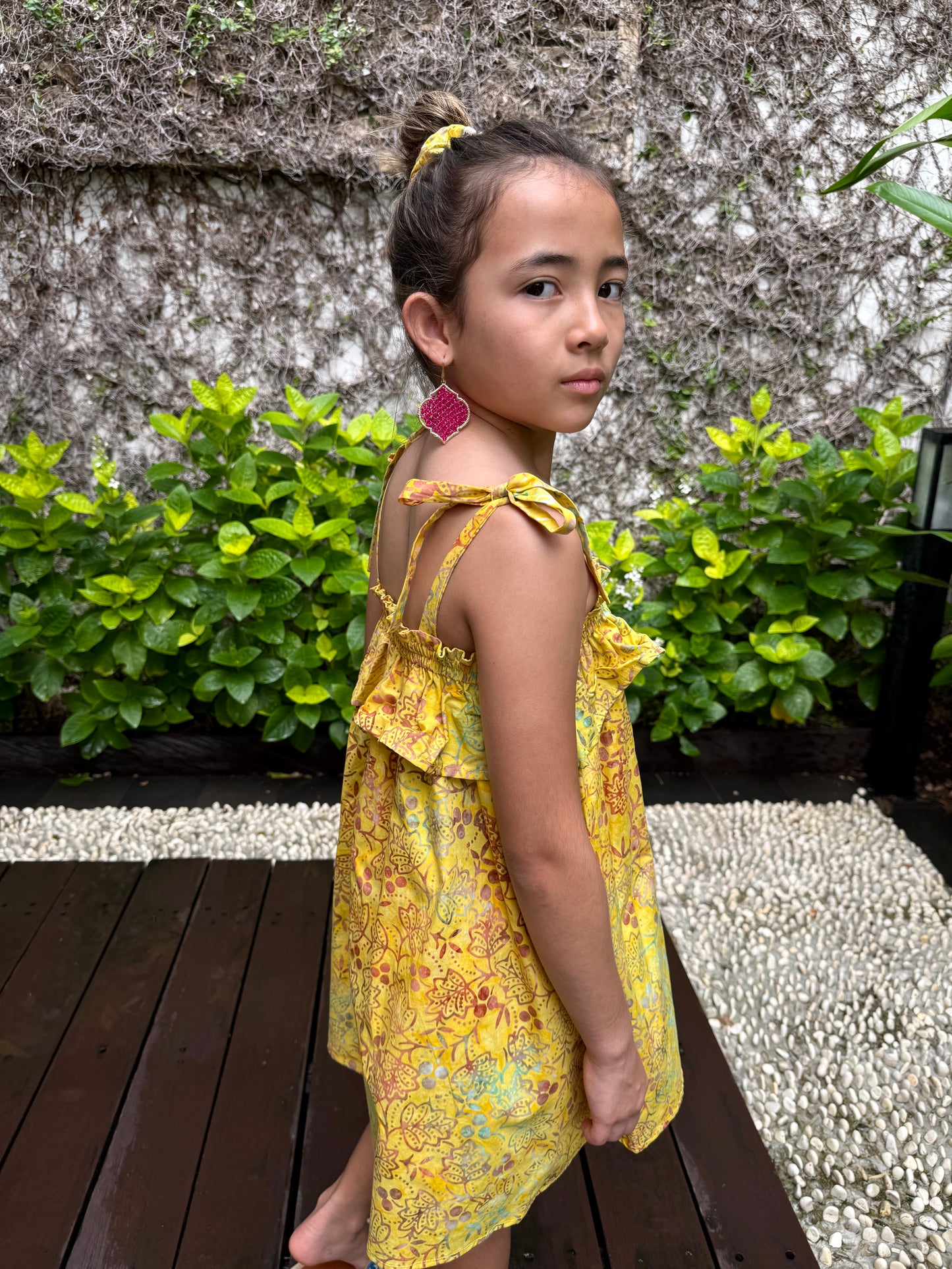 Sideways back close up view of girl wearing mustard yellow floral batik cotton sundress with adjustable shoulder ties and ruffles, sustainably made in Bali, available in Dubai