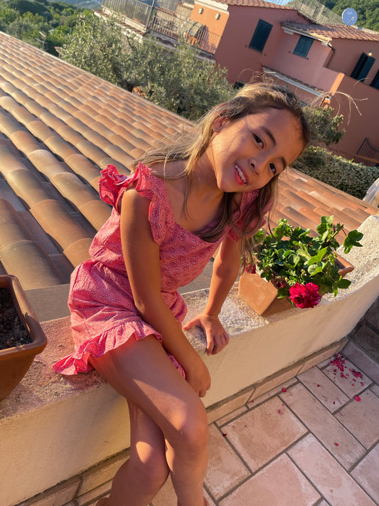 A child wearing a rose pink kids coord set with a floral block print design, sitting on a balcony with a plant pot in the foreground.