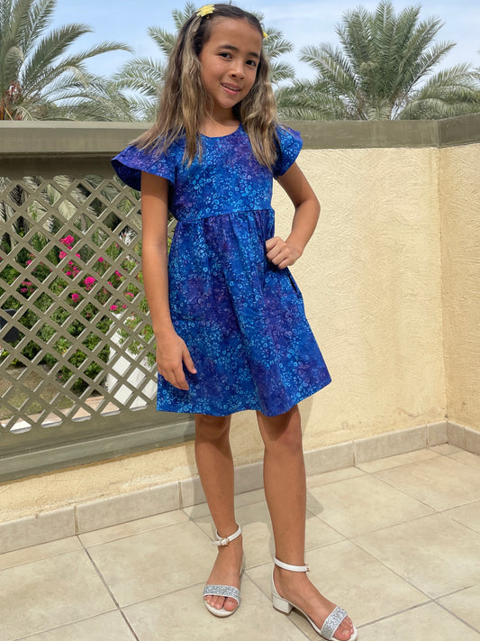 Front view of girl wearing a cotton batik dress in a contrasting blue floral print, showing the ruffle sleeves and a-line shape of the dress, sustainably handmade in Bali, for sale in Dubai.