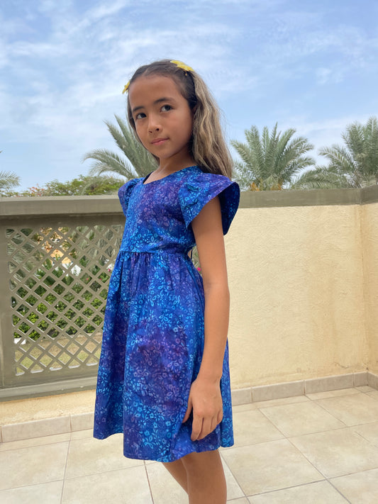 Side view of girl wearing a cotton batik dress in a contrasting blue floral print, showing the ruffle sleeves and a-line shape of the dress, sustainably handmade in Bali, for sale in Dubai.