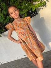 Load image into Gallery viewer, Girl wearing the Ivy orange tiger lily cotton batik dress, handmade sustainably in Bali by local artisans, featuring ruffle sleeves, adjustable back ties, and an a-line fit, perfect for outings in Dubai and those who love slow fashion.
