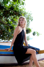 Load image into Gallery viewer, A side angle of our model sitting while wearing our Mimi black midi dress, highlighting the adjustable neck ties and graceful flow of the fabric.
