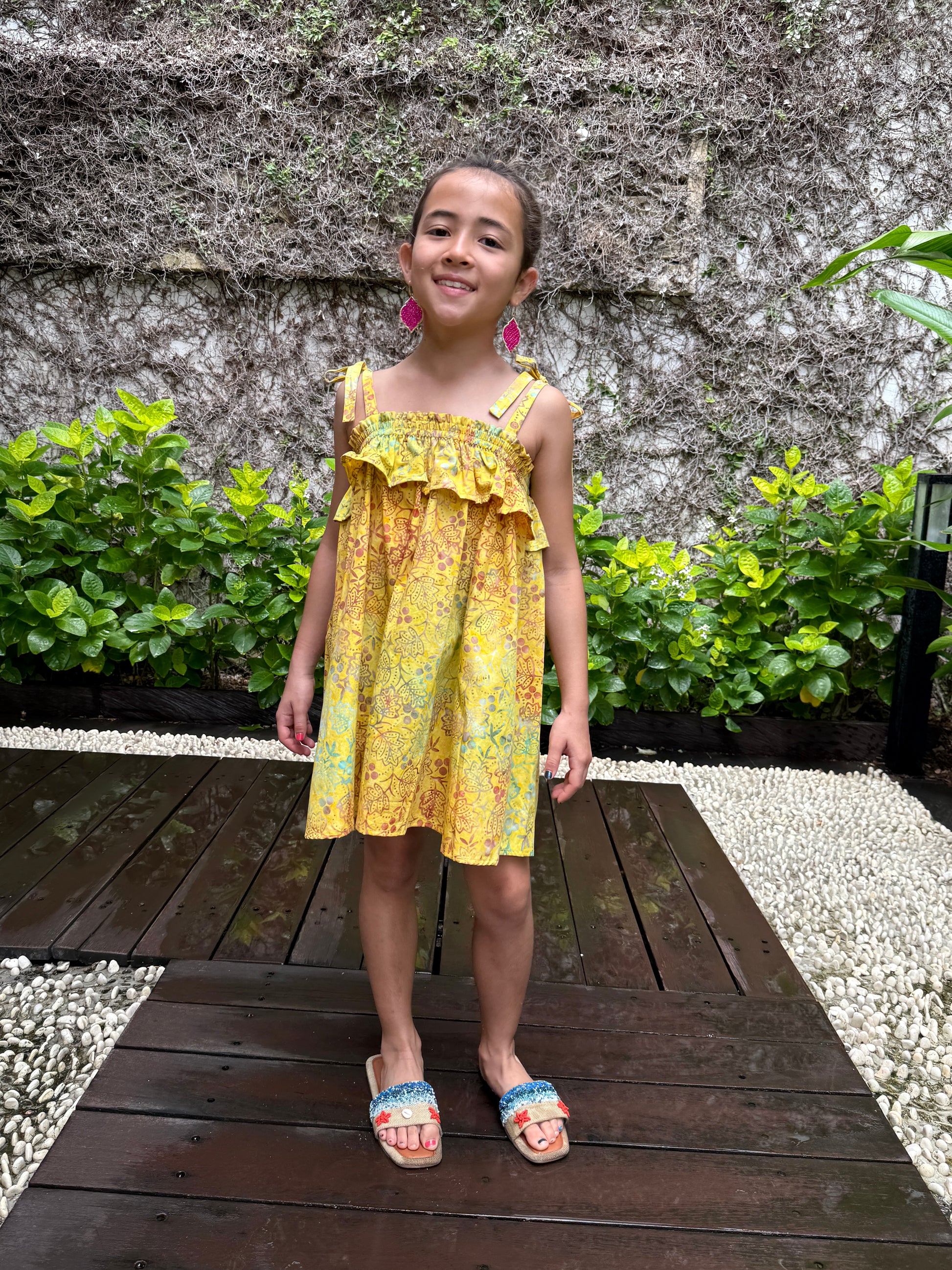 Front pose of girl wearing mustard yellow floral batik cotton sundress with adjustable shoulder ties and ruffles, sustainably made in Bali, available in Dubai