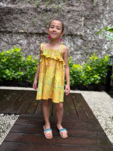 Load image into Gallery viewer, Front pose of girl wearing mustard yellow floral batik cotton sundress with adjustable shoulder ties and ruffles, sustainably made in Bali, available in Dubai