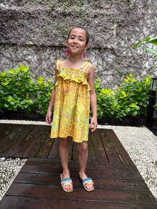 Front pose of girl wearing mustard yellow floral batik cotton sundress with adjustable shoulder ties and ruffles, sustainably made in Bali, available in Dubai