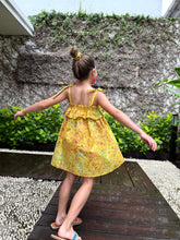 Load image into Gallery viewer, Girl twirling and wearing mustard yellow floral batik cotton sundress with adjustable shoulder ties and ruffles in a back pose