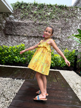 Load image into Gallery viewer, Girl wearing mustard yellow floral batik cotton sundress with adjustable shoulder ties and ruffles in a side pose