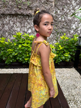 Load image into Gallery viewer, Sideways back close up view of girl wearing mustard yellow floral batik cotton sundress with adjustable shoulder ties and ruffles, sustainably made in Bali, available in Dubai
