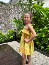 Load image into Gallery viewer, Side pose of girl wearing mustard yellow floral batik cotton sundress with adjustable shoulder ties and ruffles, sustainably made in Bali, available in Dubai
